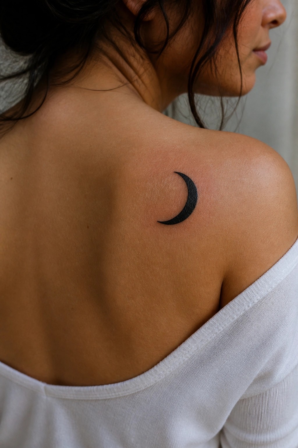 Small solid black crescent moon tattoo on a woman's upper shoulder