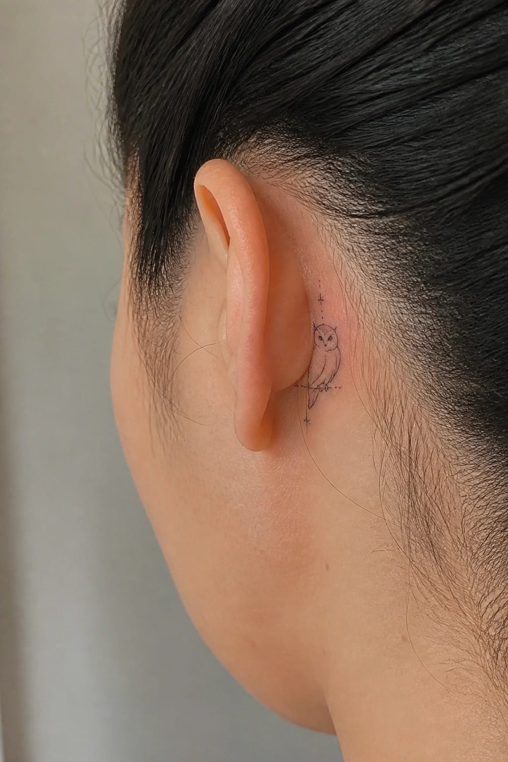 Small fine line owl tattoo placed behind the ear with simple dotted accents around it.