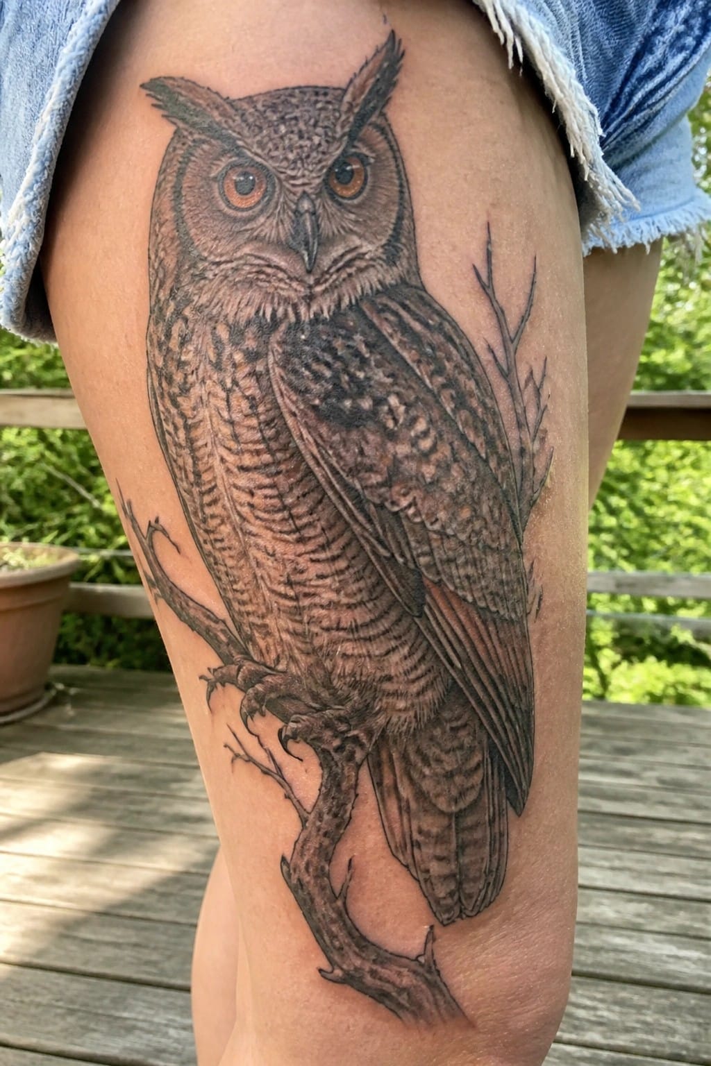 Realistic black and gray owl tattoo on a thigh, perched on a branch with detailed feathers and orange eyes.