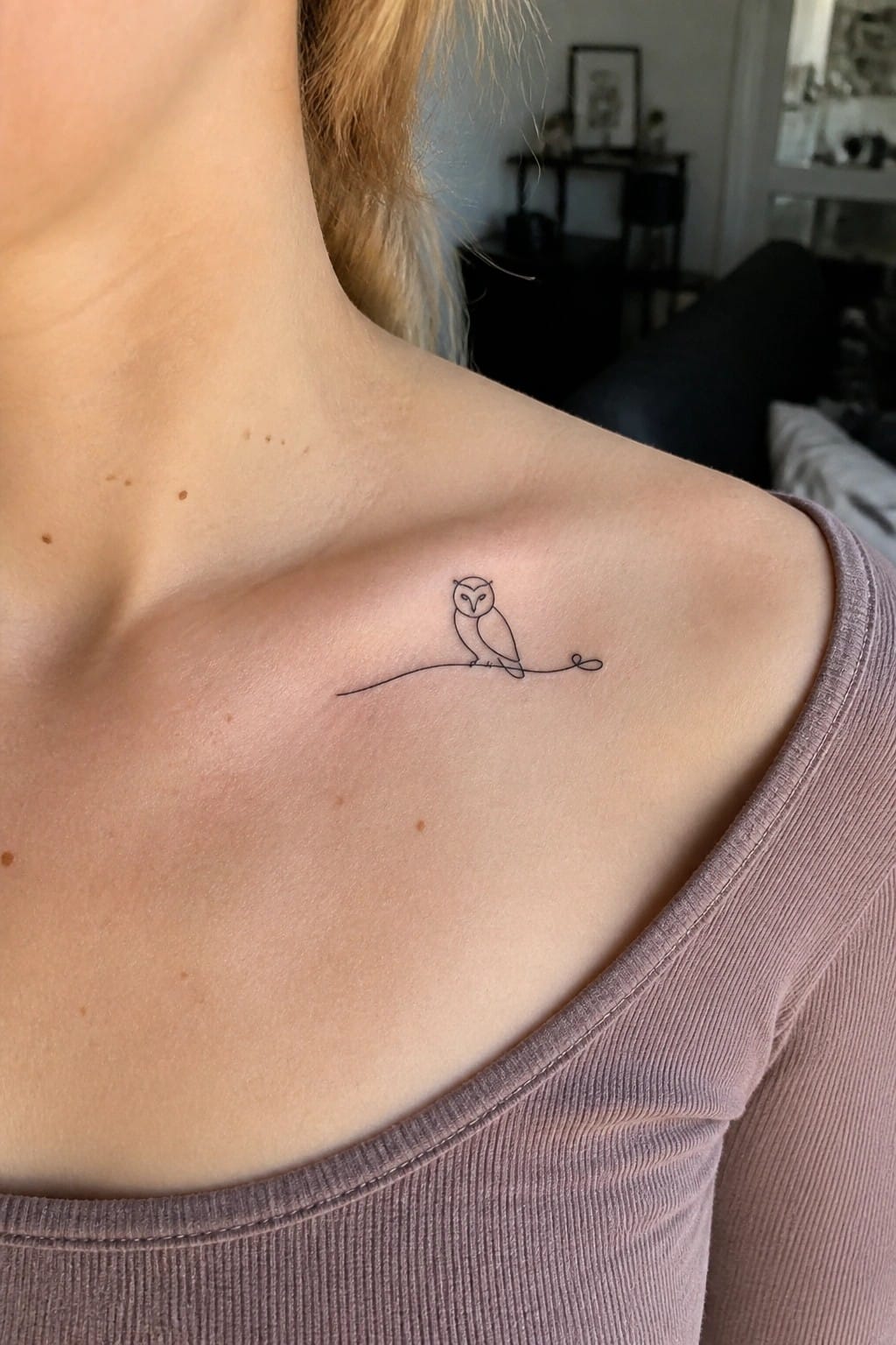 Minimalist black owl tattoo on freckled shoulder above a purple top