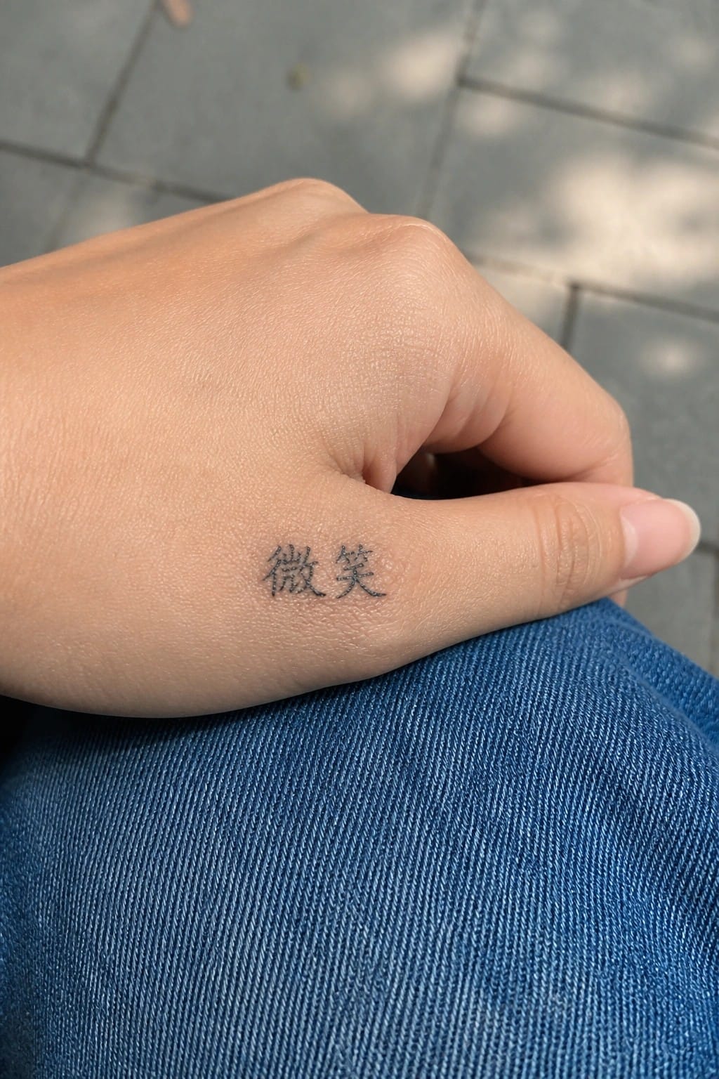 Hand with black Chinese tattoo resting on blue denim jeans over pavement.