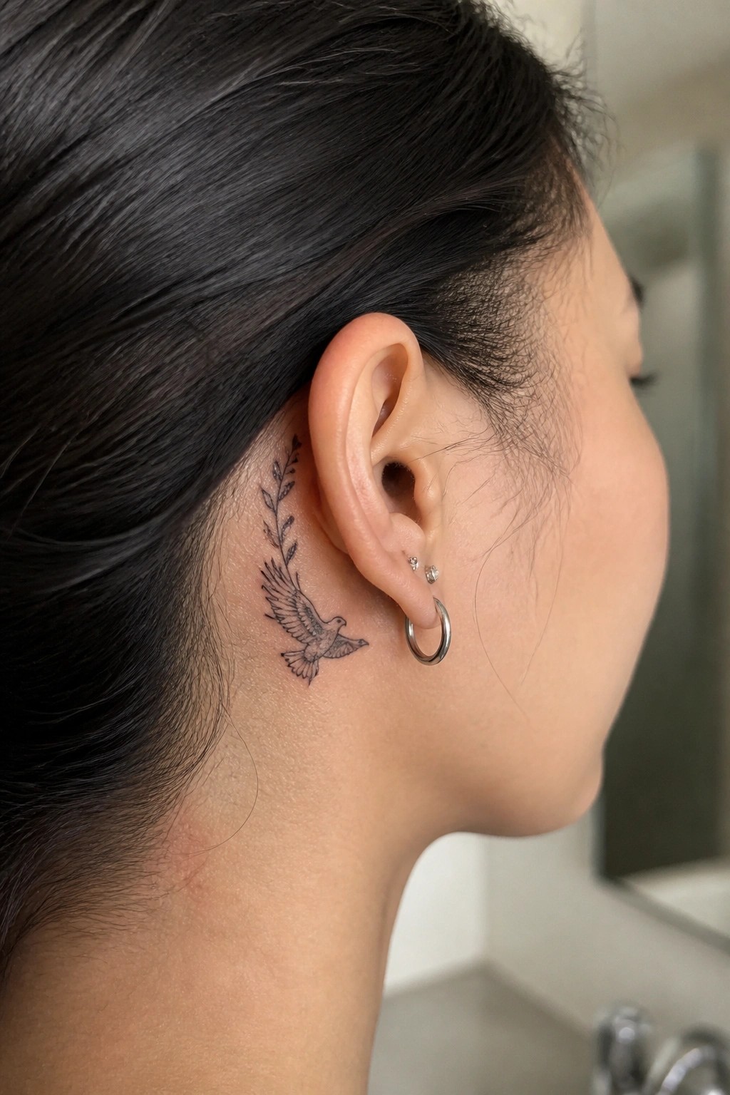 Fine line tattoo of a flying bird with a leafy branch positioned behind a woman's ear on the neck.