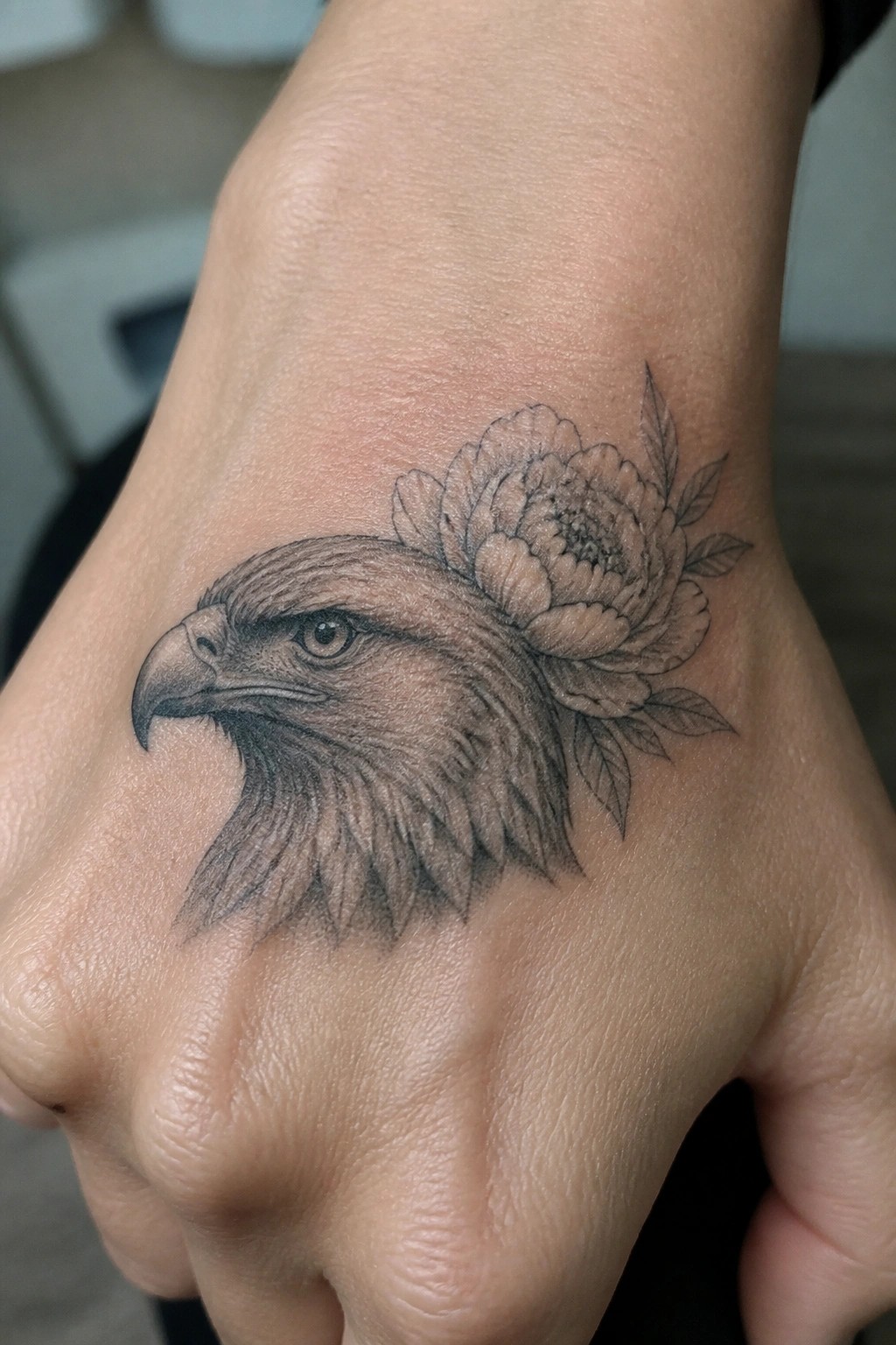 Detailed black-and-grey eagle head and peony tattoo on a clenched hand.