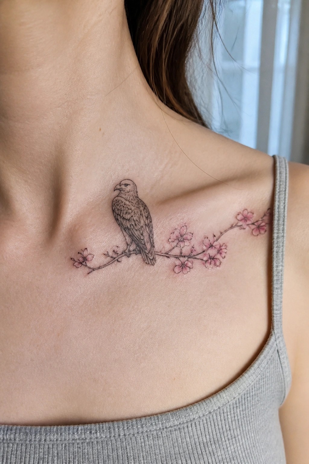 Eagle tattoo on pink cherry blossom branch across collarbone over gray tank top.