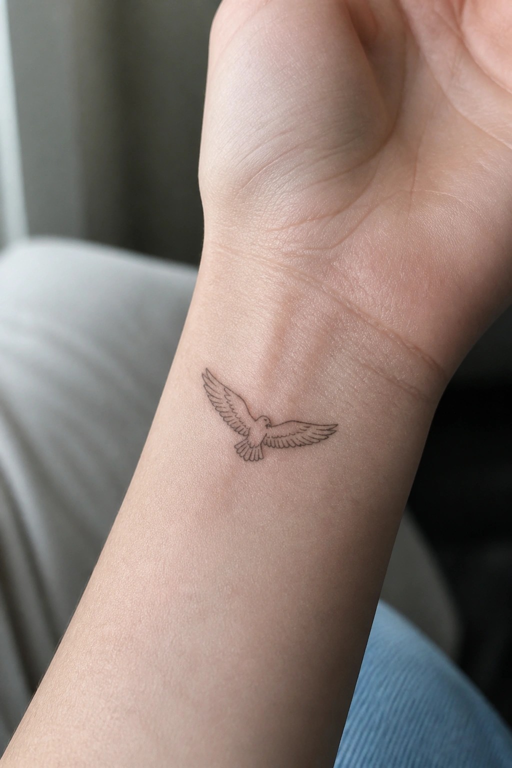 Small black bird tattoo with spread wings on light-skinned wrist near blue jeans