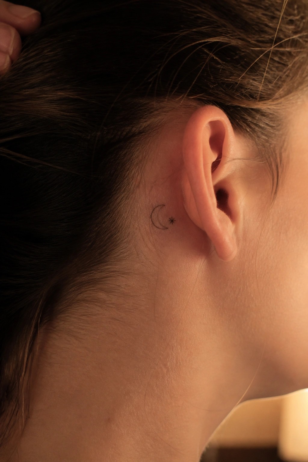 Close-up of crescent moon and star tattoo behind a person's ear with brown hair