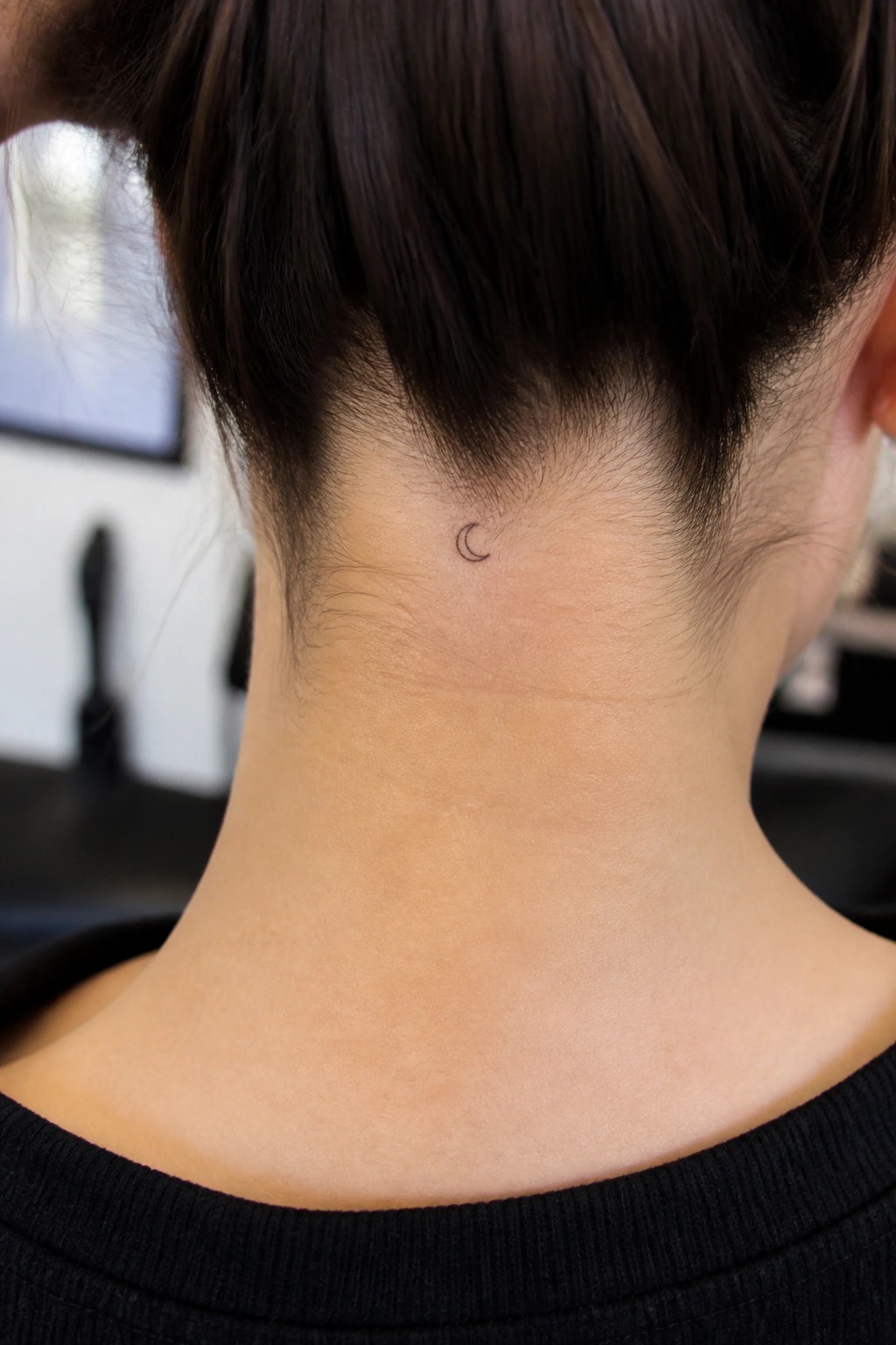 Nape of neck with small crescent moon tattoo below dark hair in a bun.