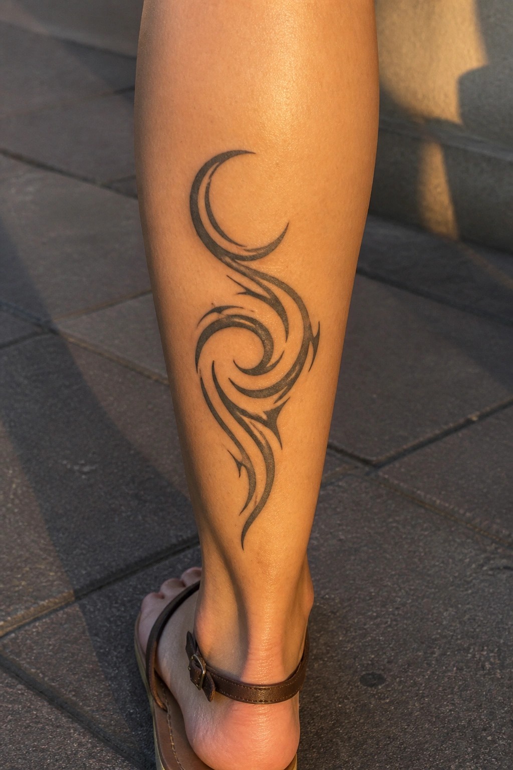 Tribal black tattoo with crescent moon and swirls on leg above brown sandal