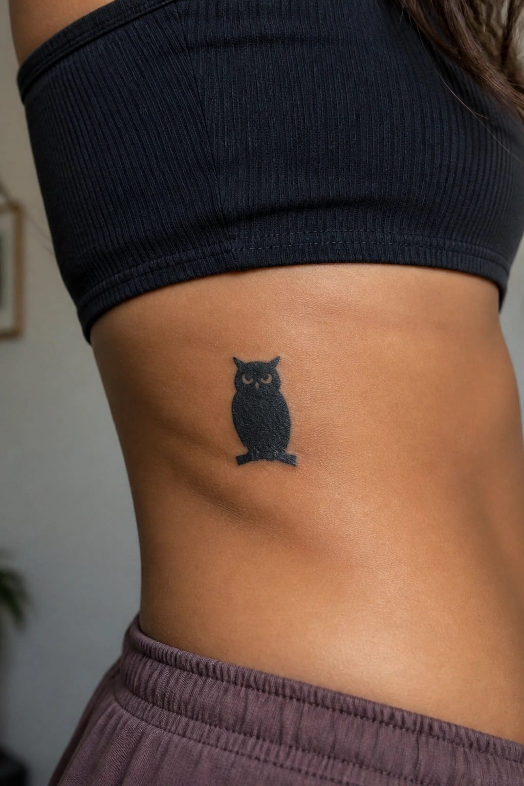 Small solid black owl tattoo on the side of a woman's torso below the ribs