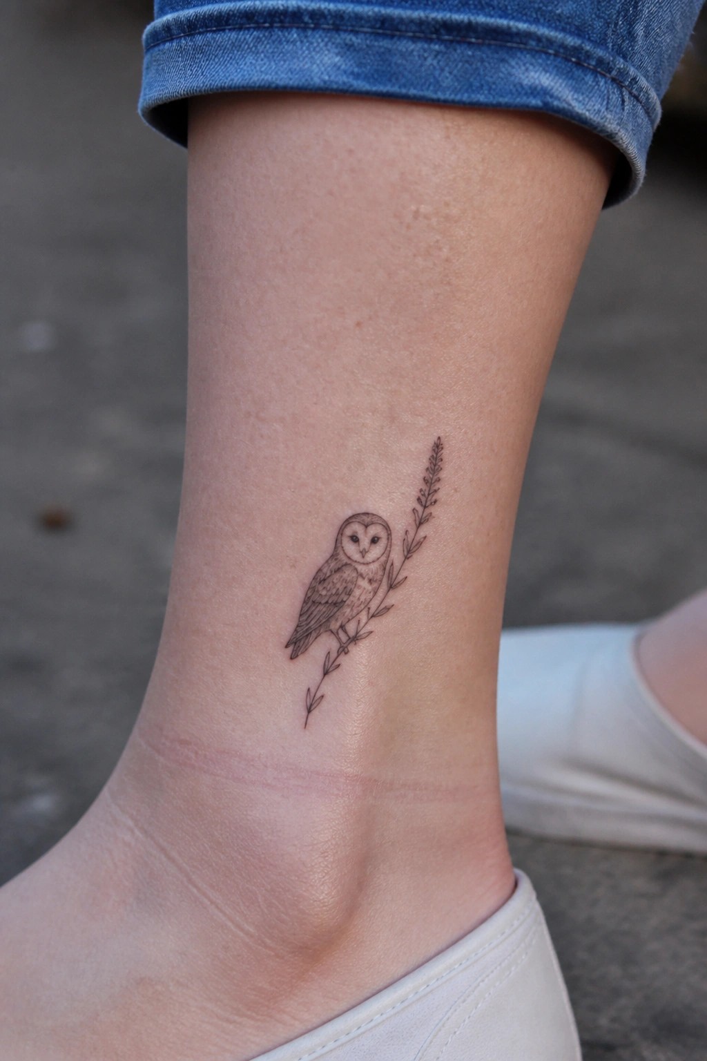 Owl tattoo on ankle beside leafy branch, below rolled blue jeans and white shoe