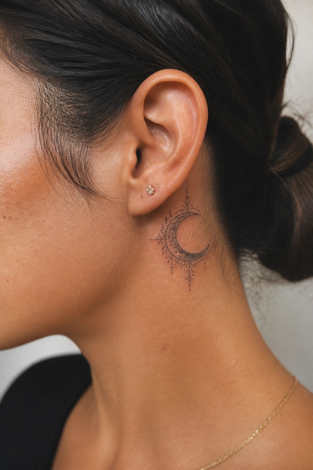 Close up of an ornamental crescent moon tattoo in fine black lines placed behind a woman's ear on the neck with small decorative details.