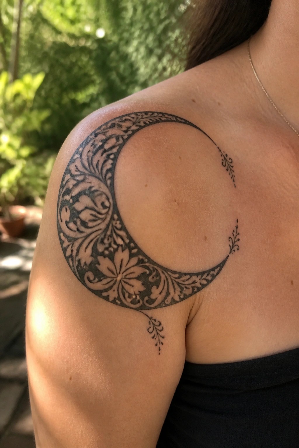 Ornate black crescent moon tattoo with florals on shoulder, black top, gold chain.