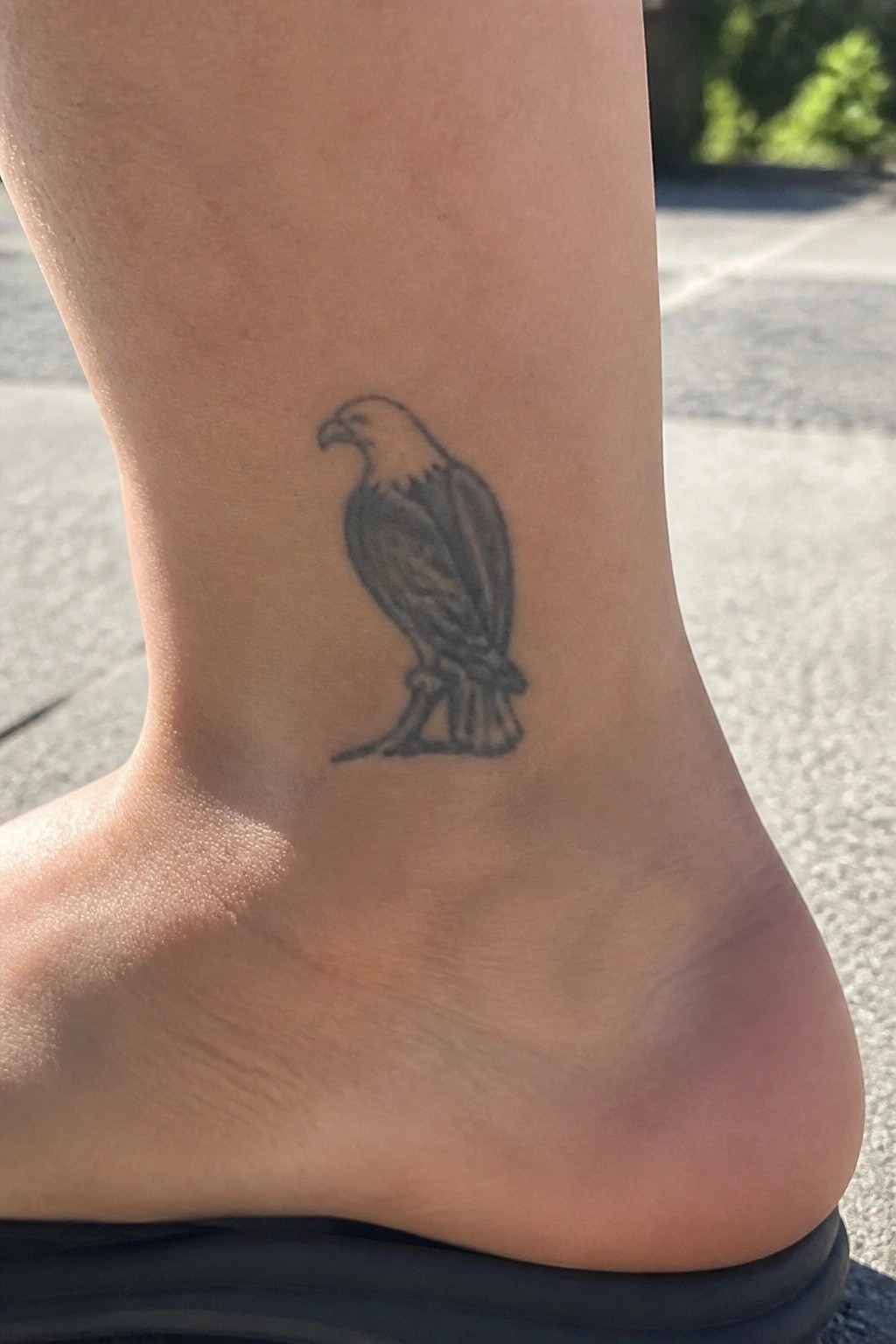 Black eagle tattoo on ankle above black sandal on pavement