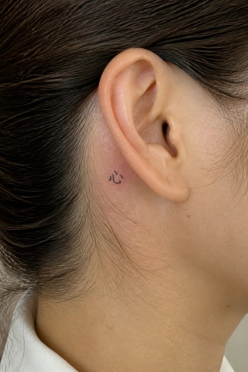 Small black heart and Chinese character tattoo behind ear on light skin.