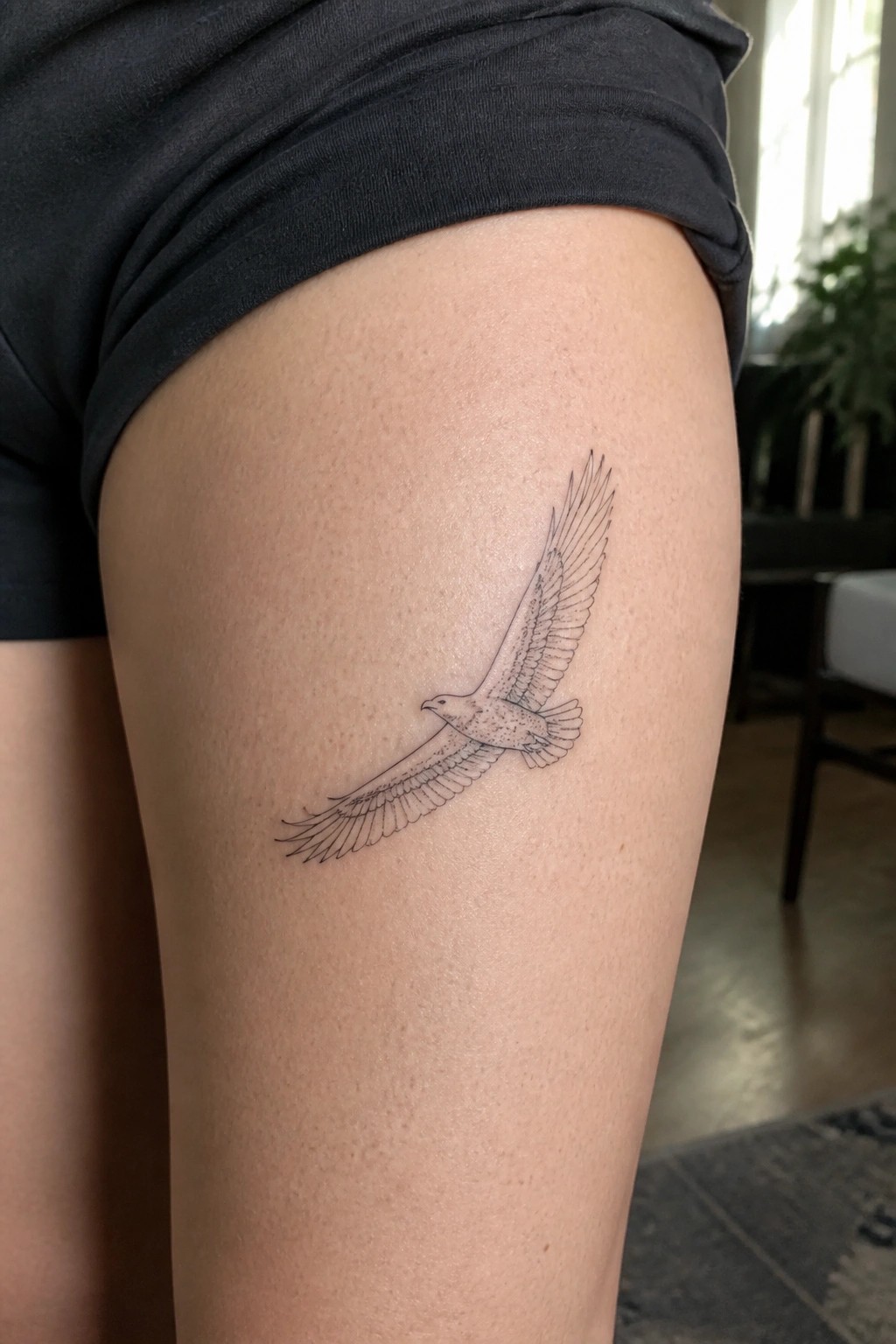 Fine-line eagle tattoo in flight on thigh beneath black shorts.
