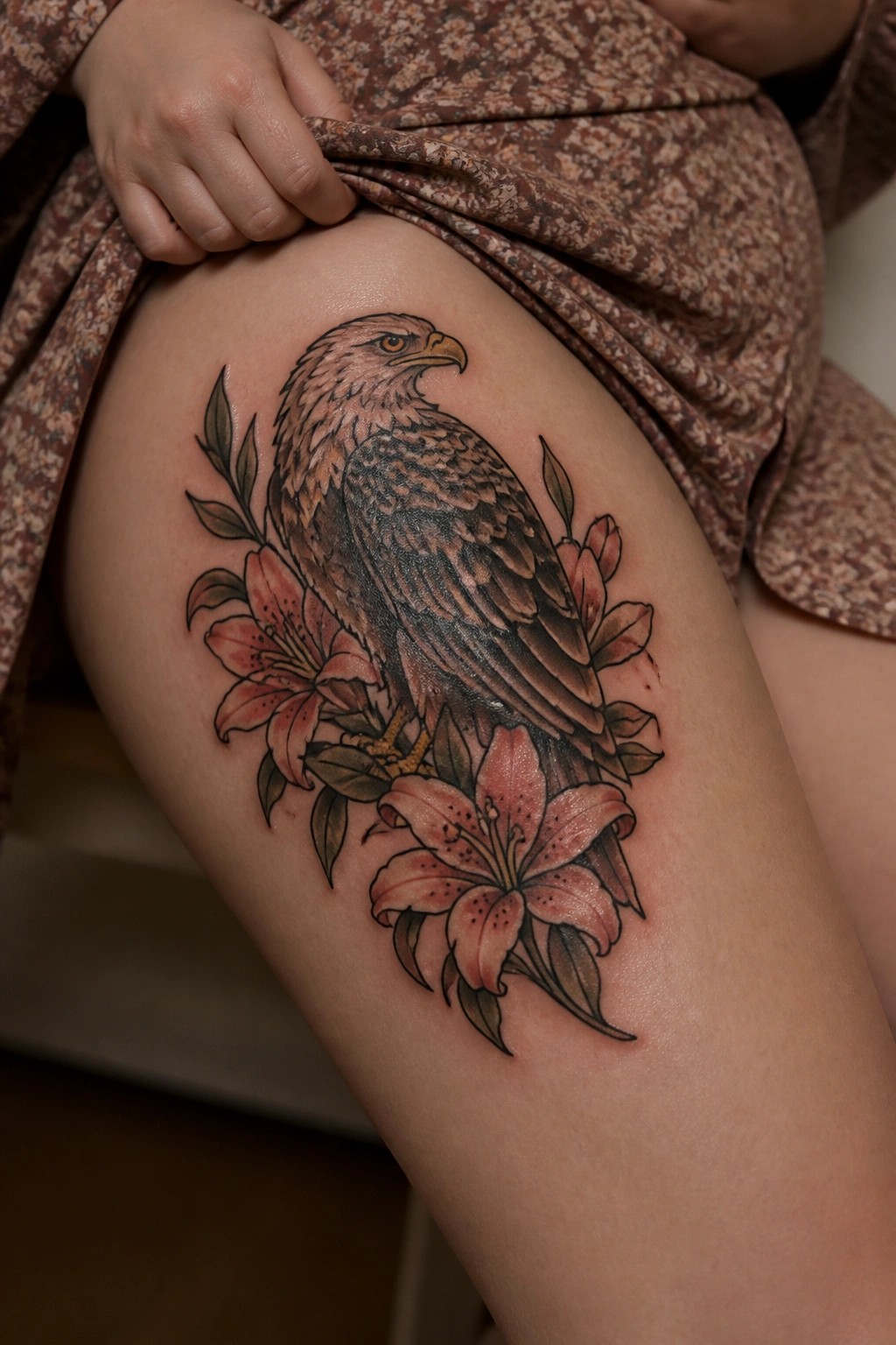 A realistic eagle tattoo on the thigh with pink lilies and leaves surrounding the base.