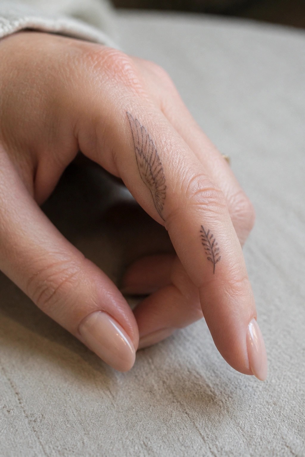 Light-skinned hand with wing tattoo on middle finger and leaf tattoo on ring finger