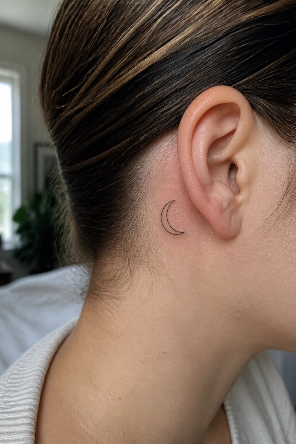 Small black outline crescent moon tattoo placed behind the ear on the side of the head