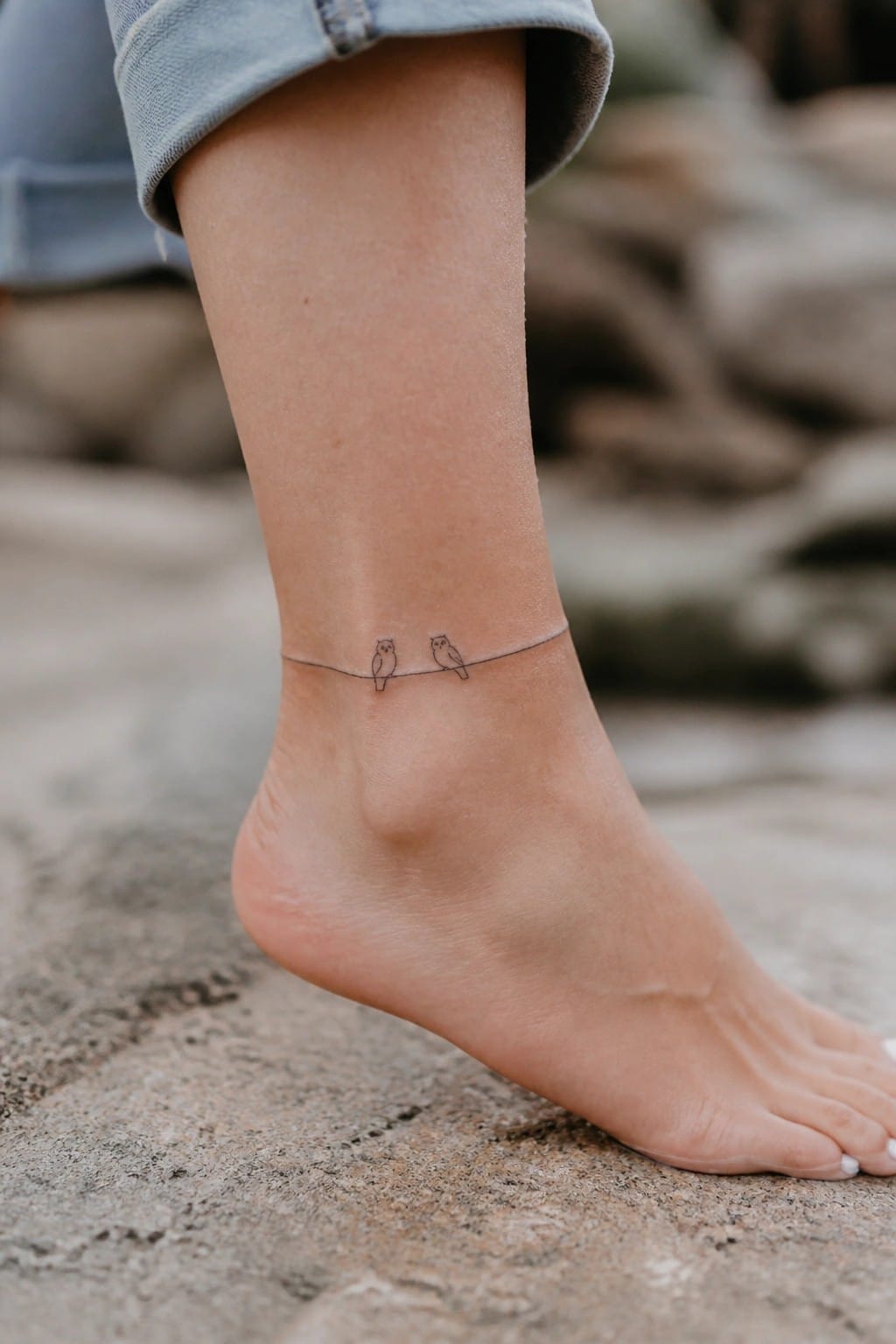 Minimalist fine line tattoo of two small owls on a thin horizontal line wrapped around an ankle.
