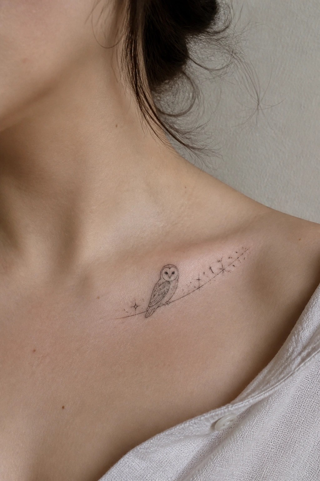 Fine line tattoo of a small owl perched on a line with stars along a woman's collarbone