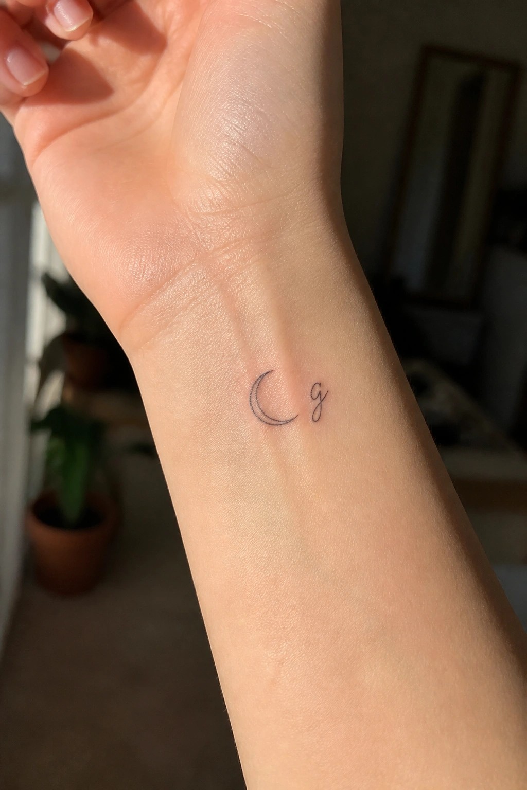 Fine line crescent moon tattoo with a small cursive initial on the inner wrist