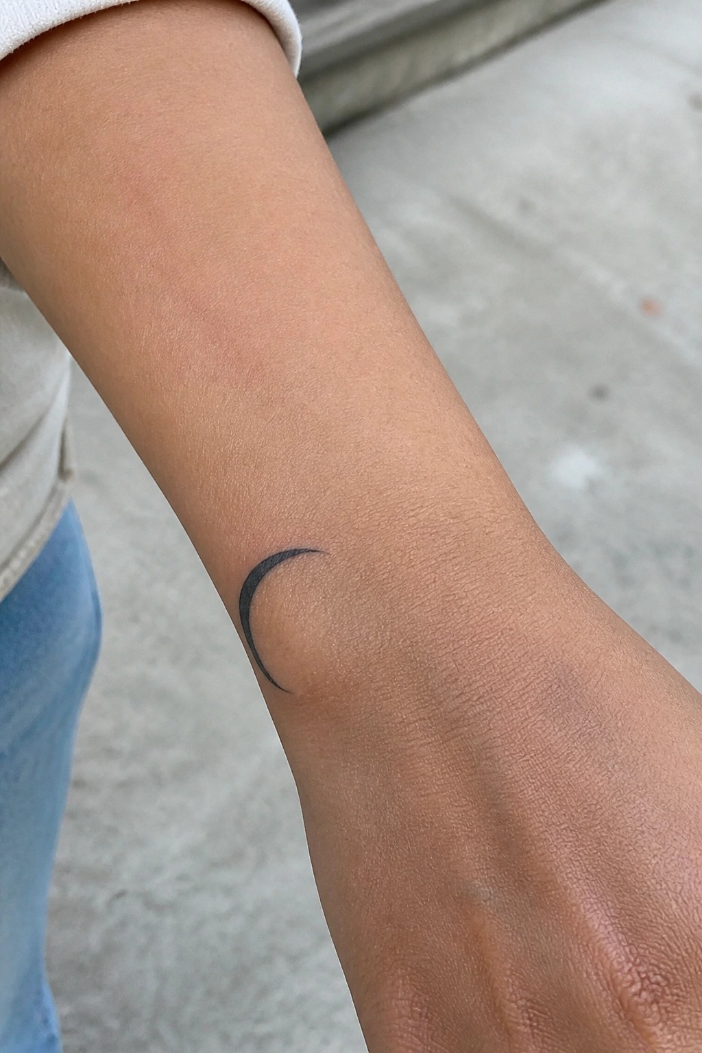 Crescent moon tattoo on wrist, white sleeve and blue jeans visible.