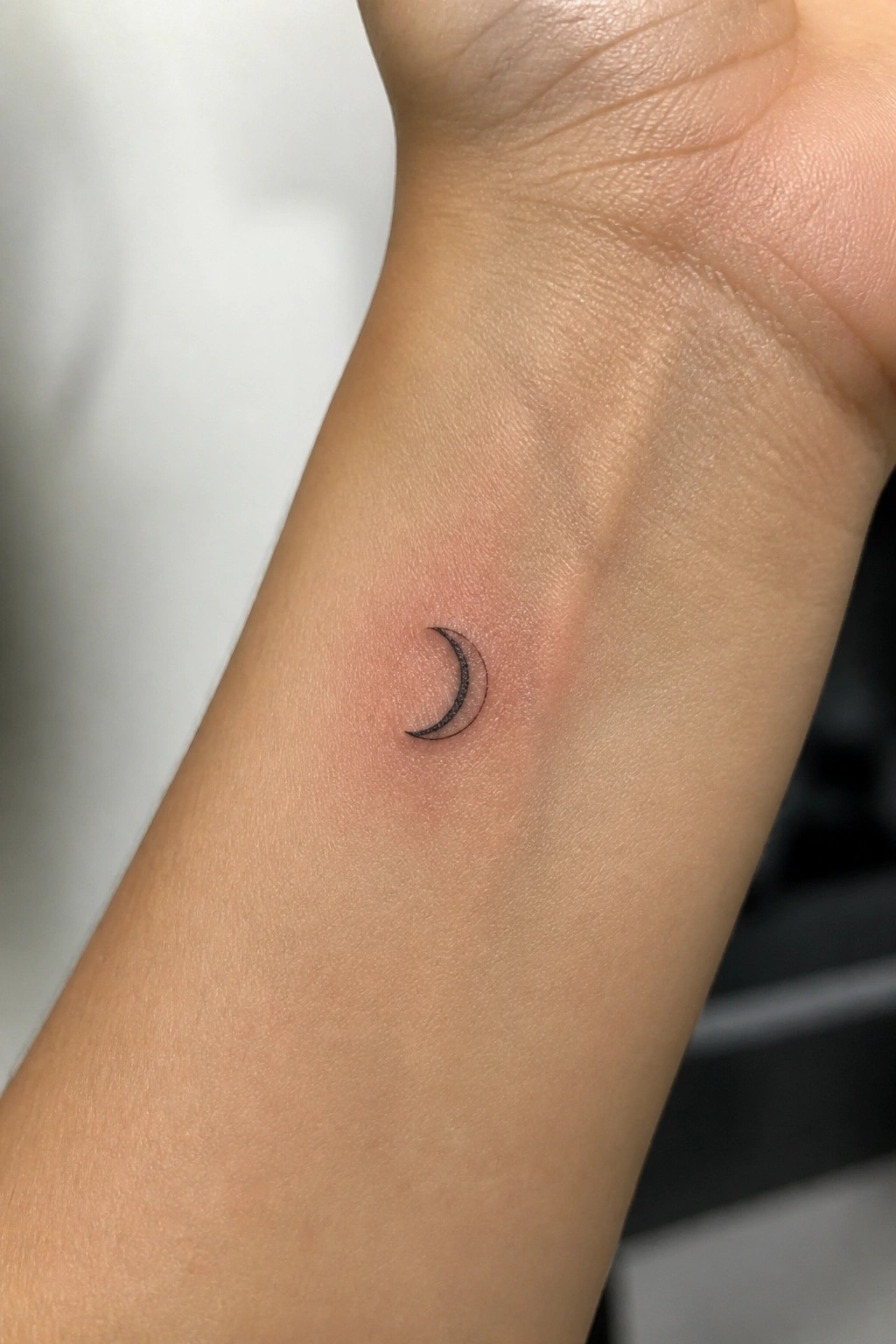 Small black crescent moon tattoo on the inner wrist with light shading in a minimal style.