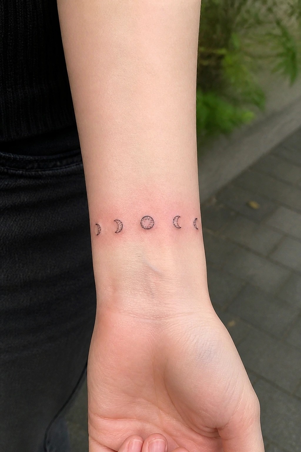 Wrist with moon phases tattoo on light skin, black sleeve visible.