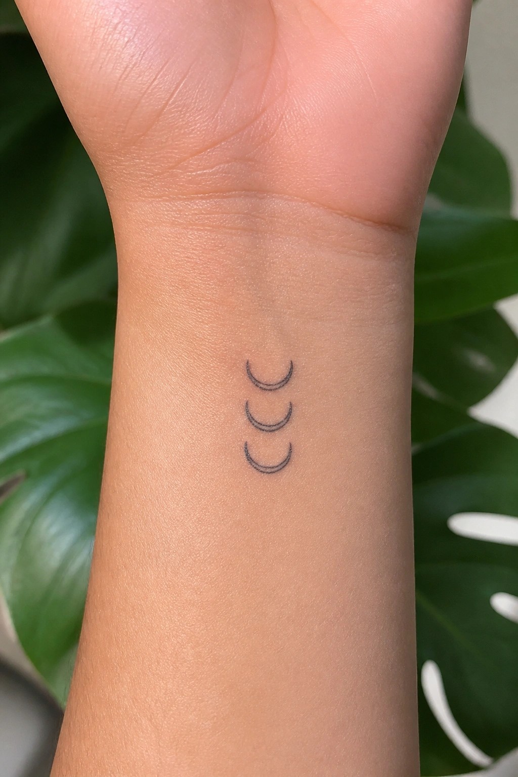 Three black crescent moon tattoos on light-skinned wrist with green leaves behind.