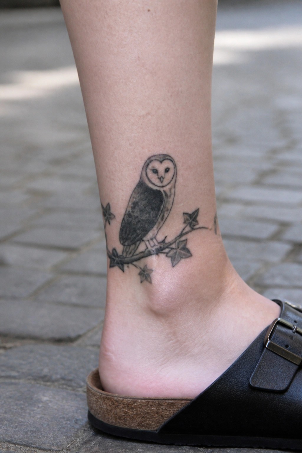 Barn owl tattoo on ankle perched on branch with leaves and stars in black and gray shading