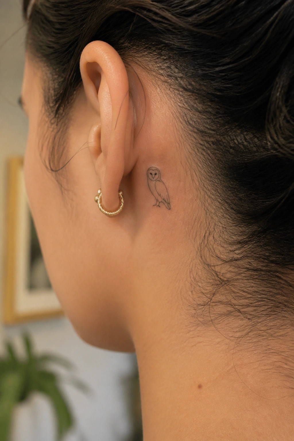 Close view of small owl tattoo behind ear with gold hoop earring