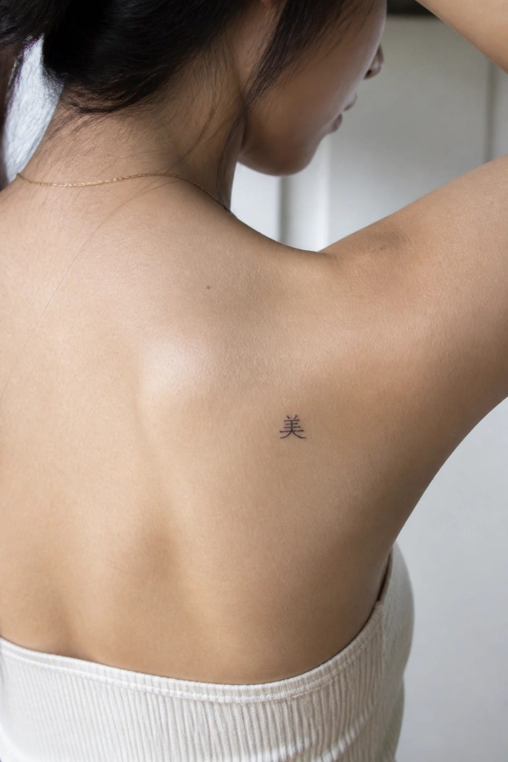 Woman's upper back with small Chinese tattoo, gold necklace, white ribbed top, dark hair in ponytail