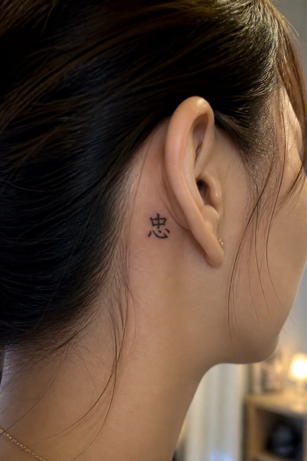 Small black Chinese character tattoo positioned behind the ear on a woman with dark hair.