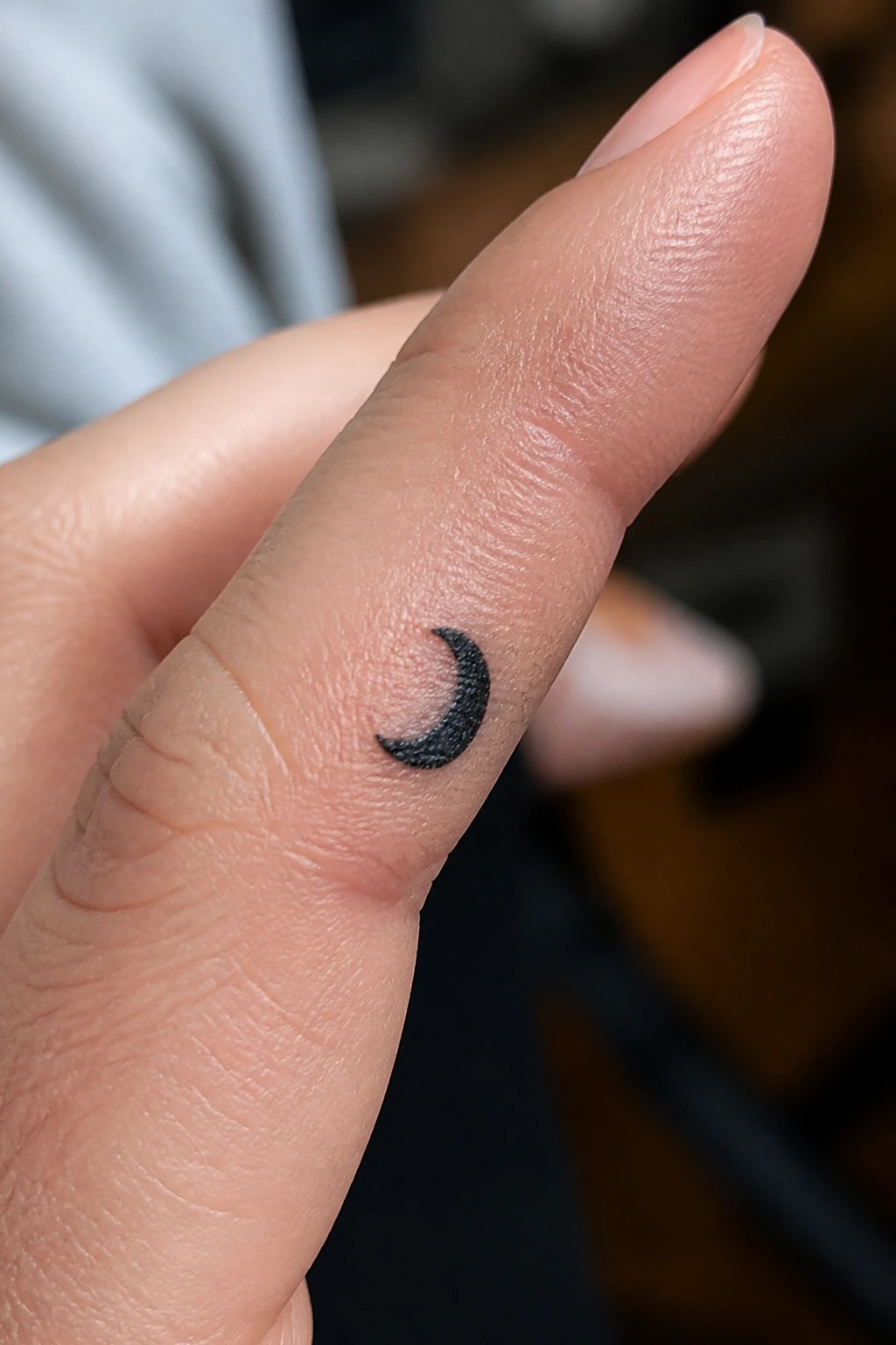 Light-skinned finger with small black crescent moon tattoo near knuckle.