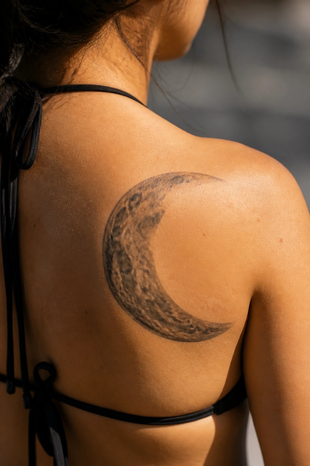 Crescent moon tattoo on tanned shoulder above black bikini top in sunlight