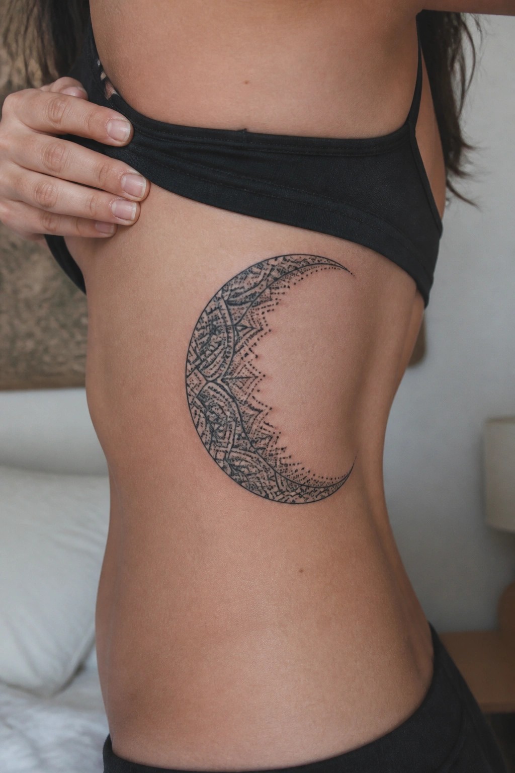Hand lifting black top to reveal ornate crescent moon tattoo on ribs