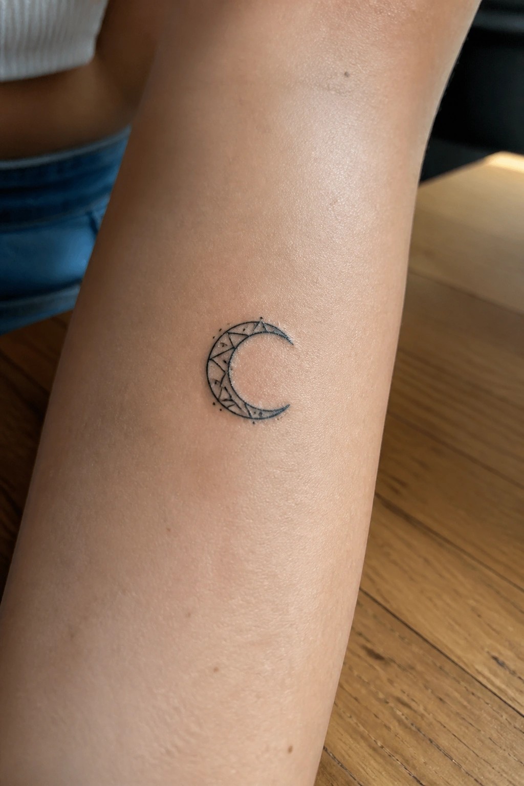 Geometric crescent moon tattoo on forearm beside white top and blue jeans on wood
