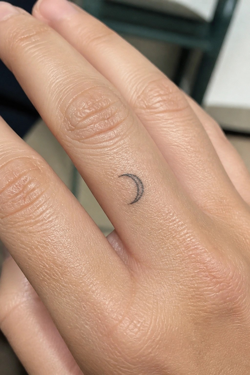 Light-skinned hand with small black crescent moon tattoo on ring finger.