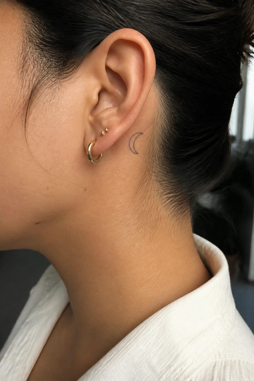 Small black crescent moon tattoo placed behind the ear on a woman's head.