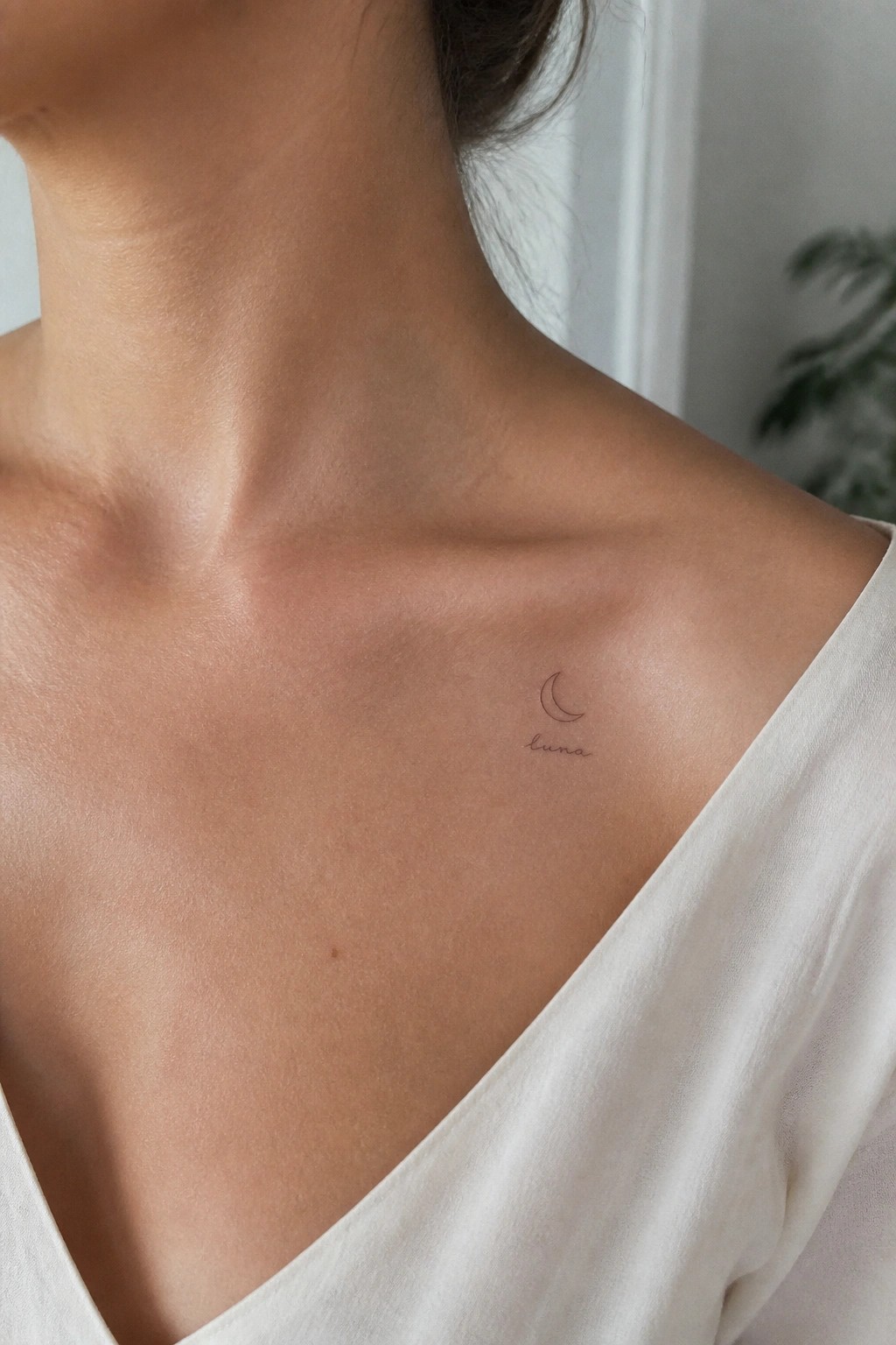 Fine line crescent moon tattoo with the word luna placed on the collarbone area of a woman wearing a white top.