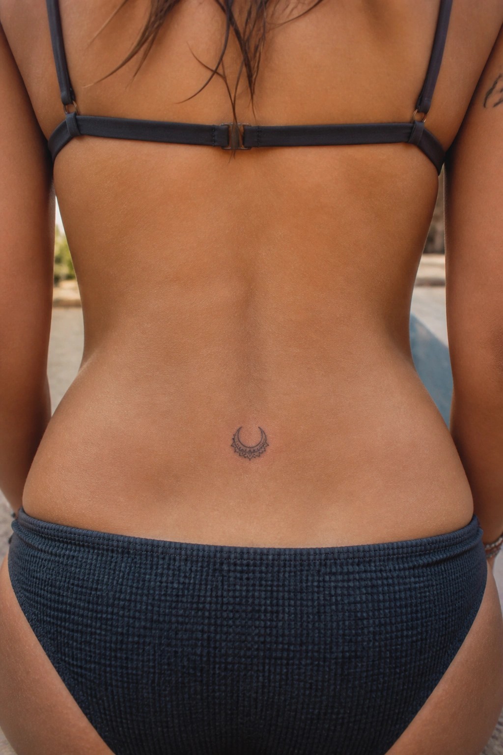 Bare tanned woman's back in dark bikini, small crescent lace tattoo on lower back.