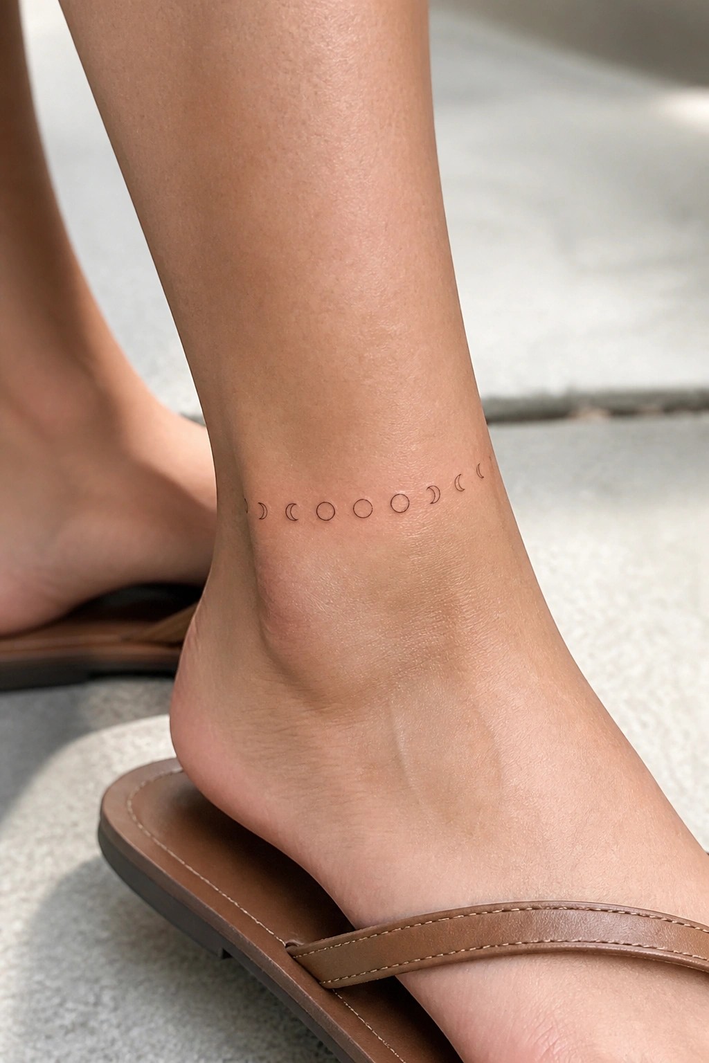 Ankle with delicate minimalist moon phase tattoo encircling ankle above tan leather flip‑flop