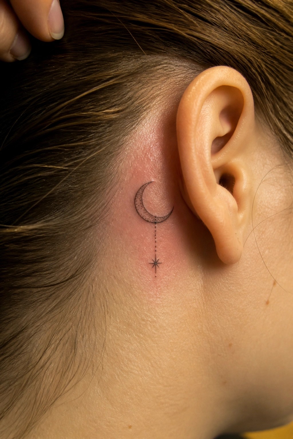Crescent moon tattoo with dotted line to star behind ear on light skin