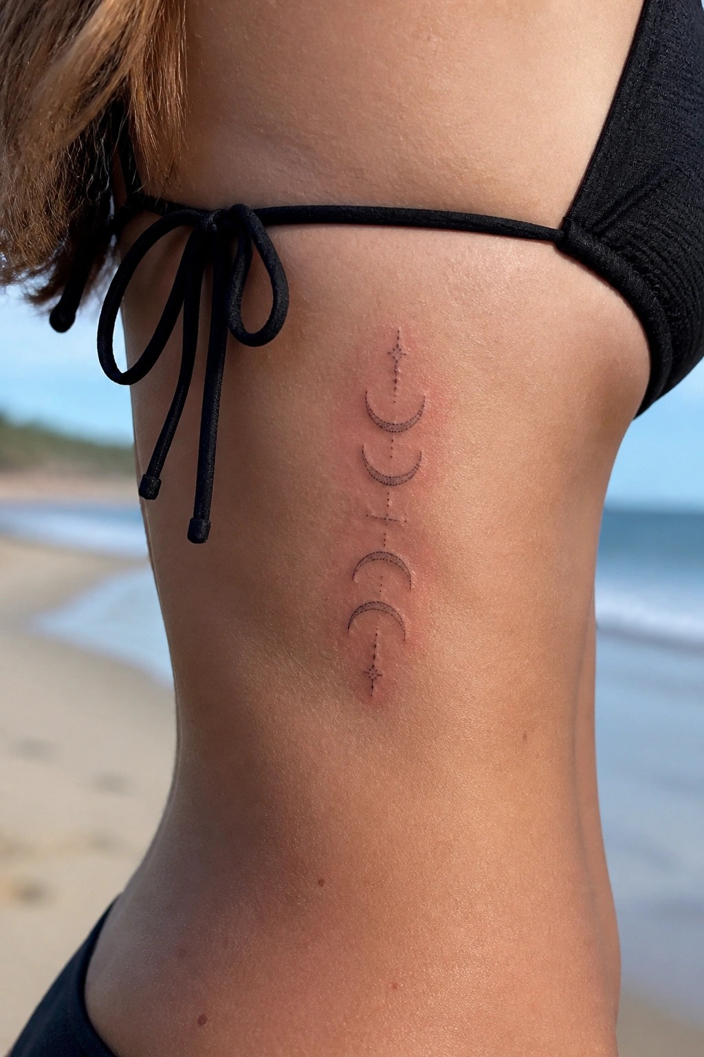 Black bikini top with vertical moon phases tattoo on side torso at beach