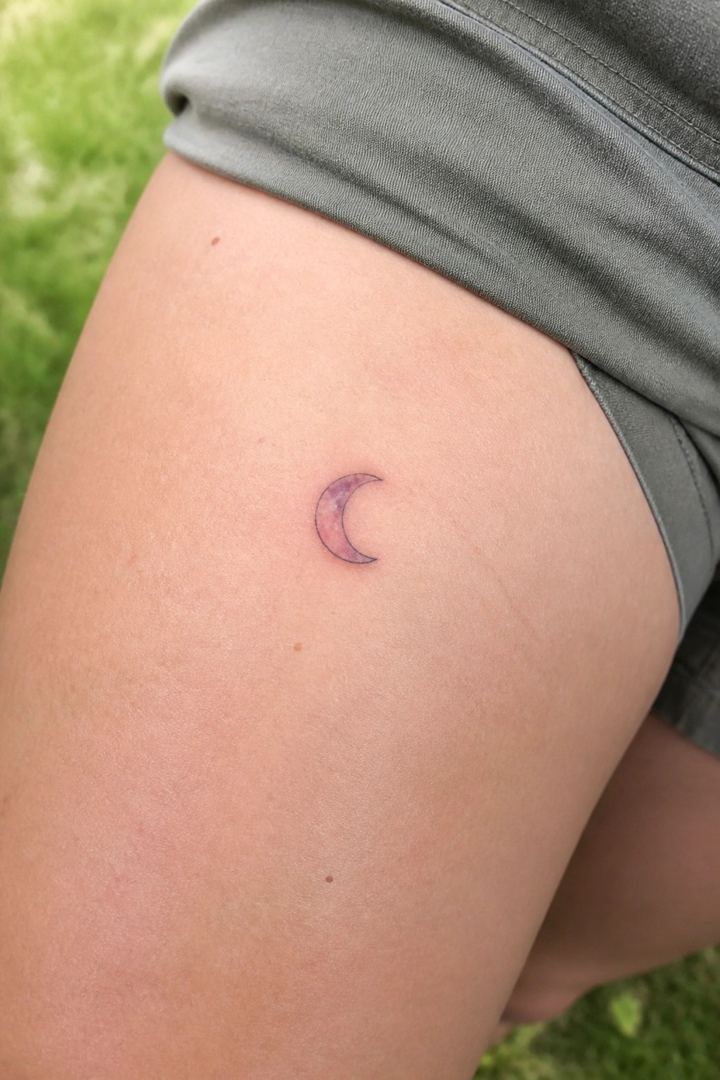 Small crescent moon tattoo with soft purple and pink watercolor shading placed on the upper thigh.