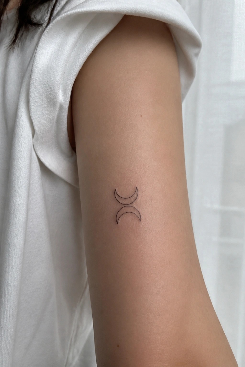 Small black line tattoo of two stacked crescent moons on the upper arm of a person wearing a white shirt.