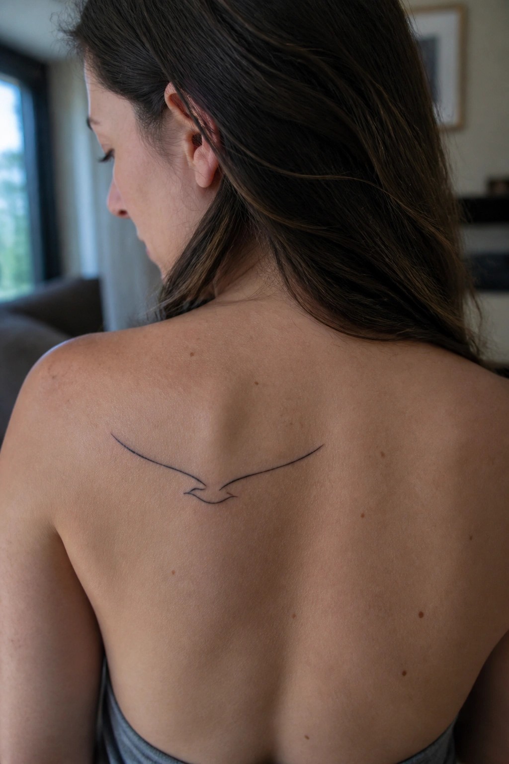Woman's bare back with minimalist bird tattoo, long brown hair over shoulder
