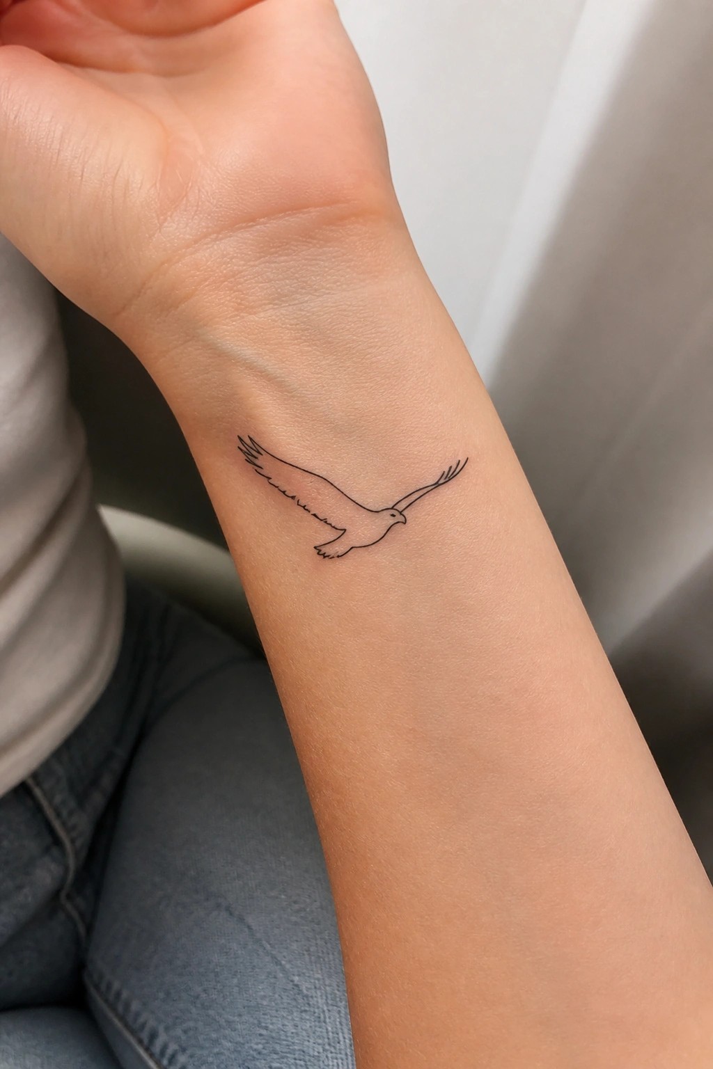 Minimalist black line tattoo of a flying bird on inner wrist above blue jeans