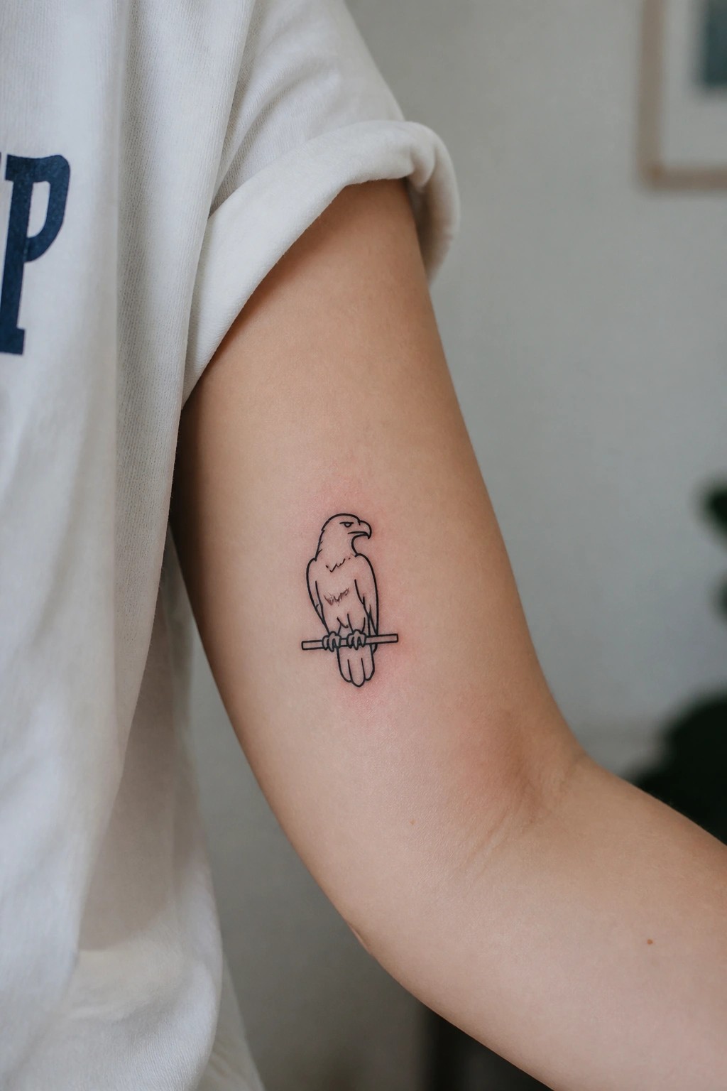 Minimalist bird tattoo on upper arm beside white t-shirt with blue lettering.
