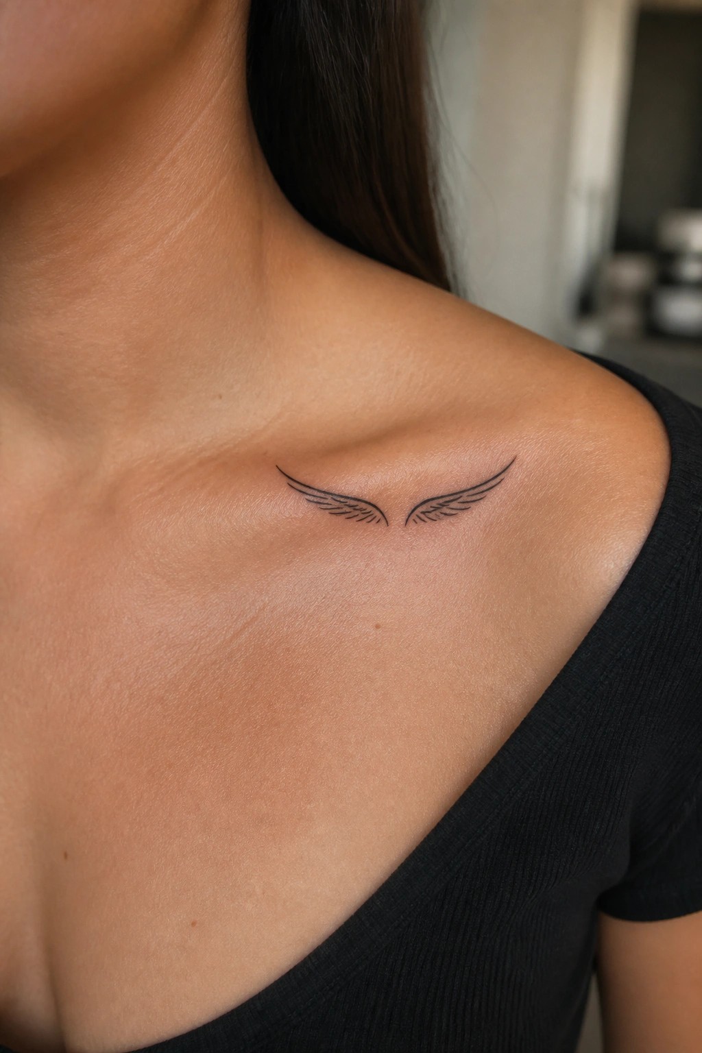 Minimalist black line art tattoo of symmetrical eagle wings placed on a woman's collarbone area